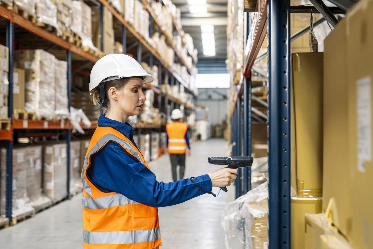 Woman worker using barcode scanner for inventory management in large logistics and distribution warehouse with racks